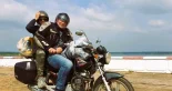 Local guide and helmeted passenger on a loaded motorbike beside the water